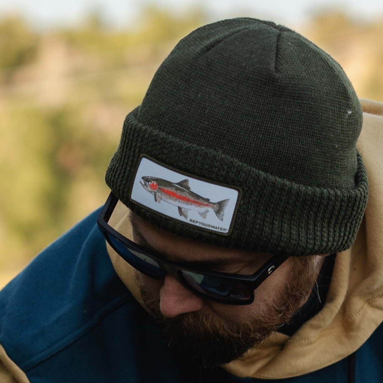 man wearing a dark green beanie with a white patch that has a rainbow trout on it 
