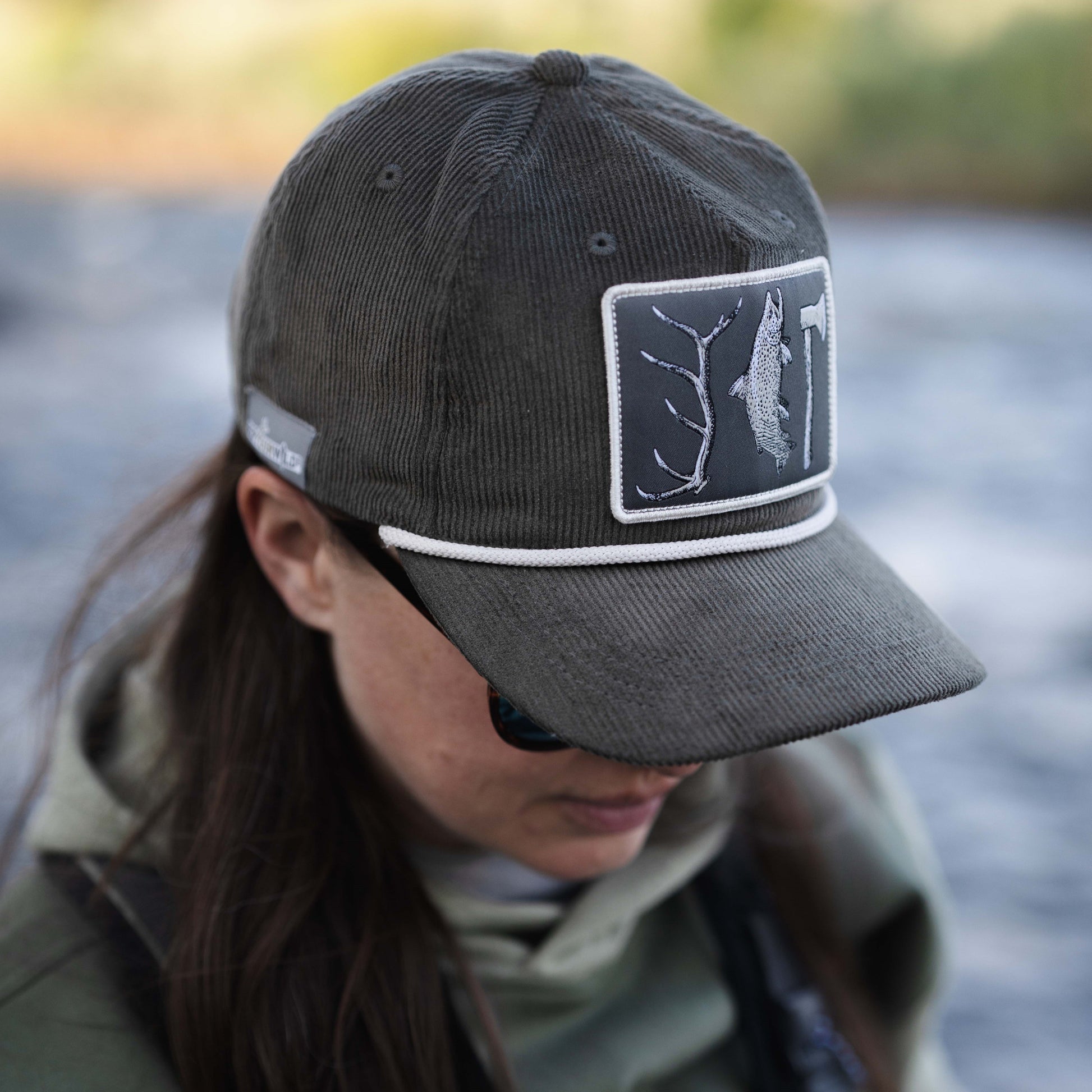 woman wearing a corduroy green hat with a white rope and a patch on the front showing a elk tine, fish, and axe pen and ink