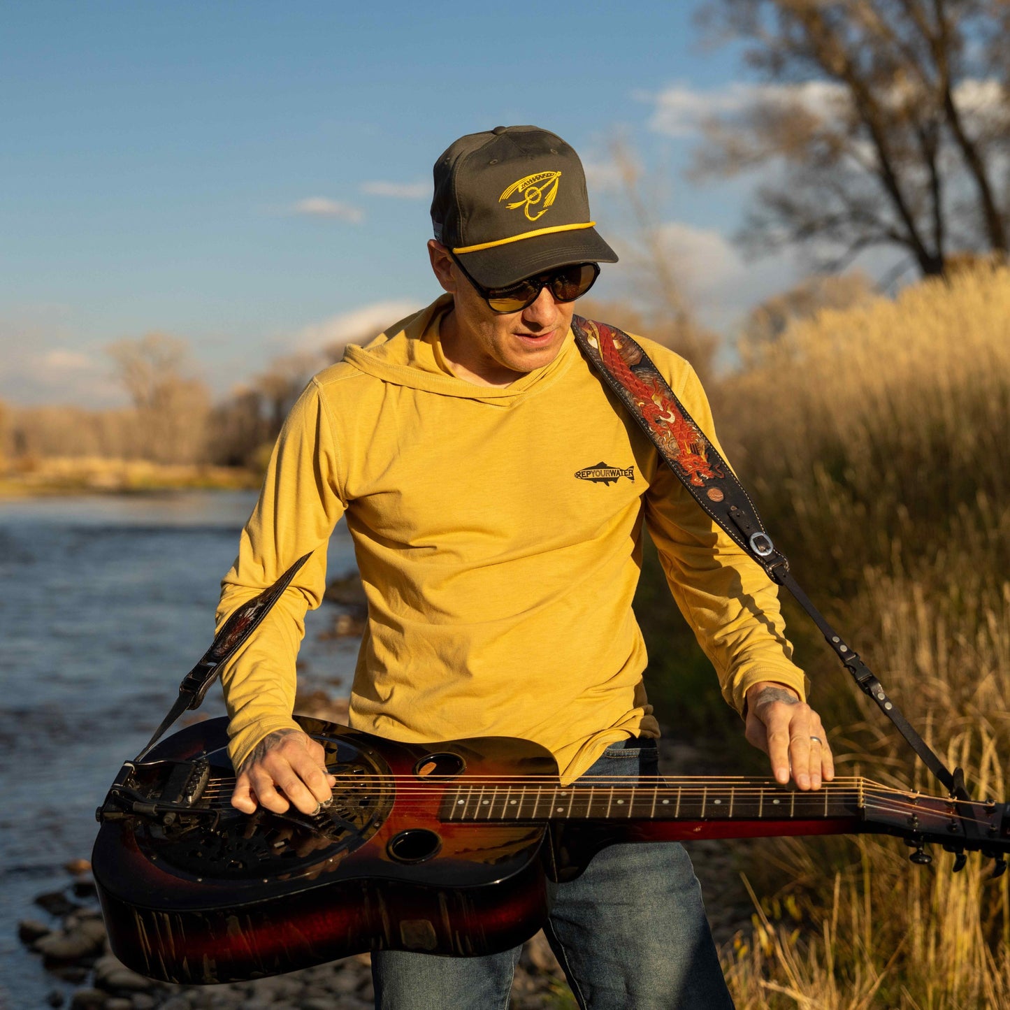 man playing guitar wearing infamous string dusters merino breeze sun hoody and unstructured 5 panel string dusters hat with banjo fly design