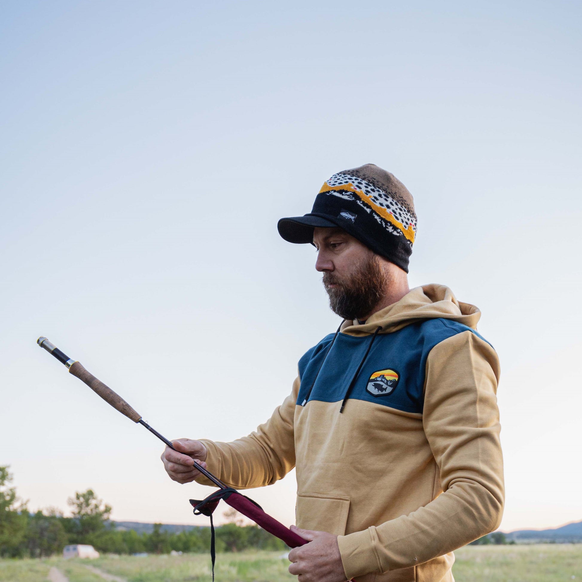 man setting up his fishing rod wearing a tan and blue sweatshirt and a brown trout beanie
