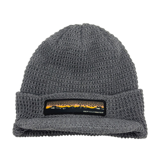 Light gray knit hat with a brim and a patch on the front showing a mountain range and brown trout skin as the "sunset"