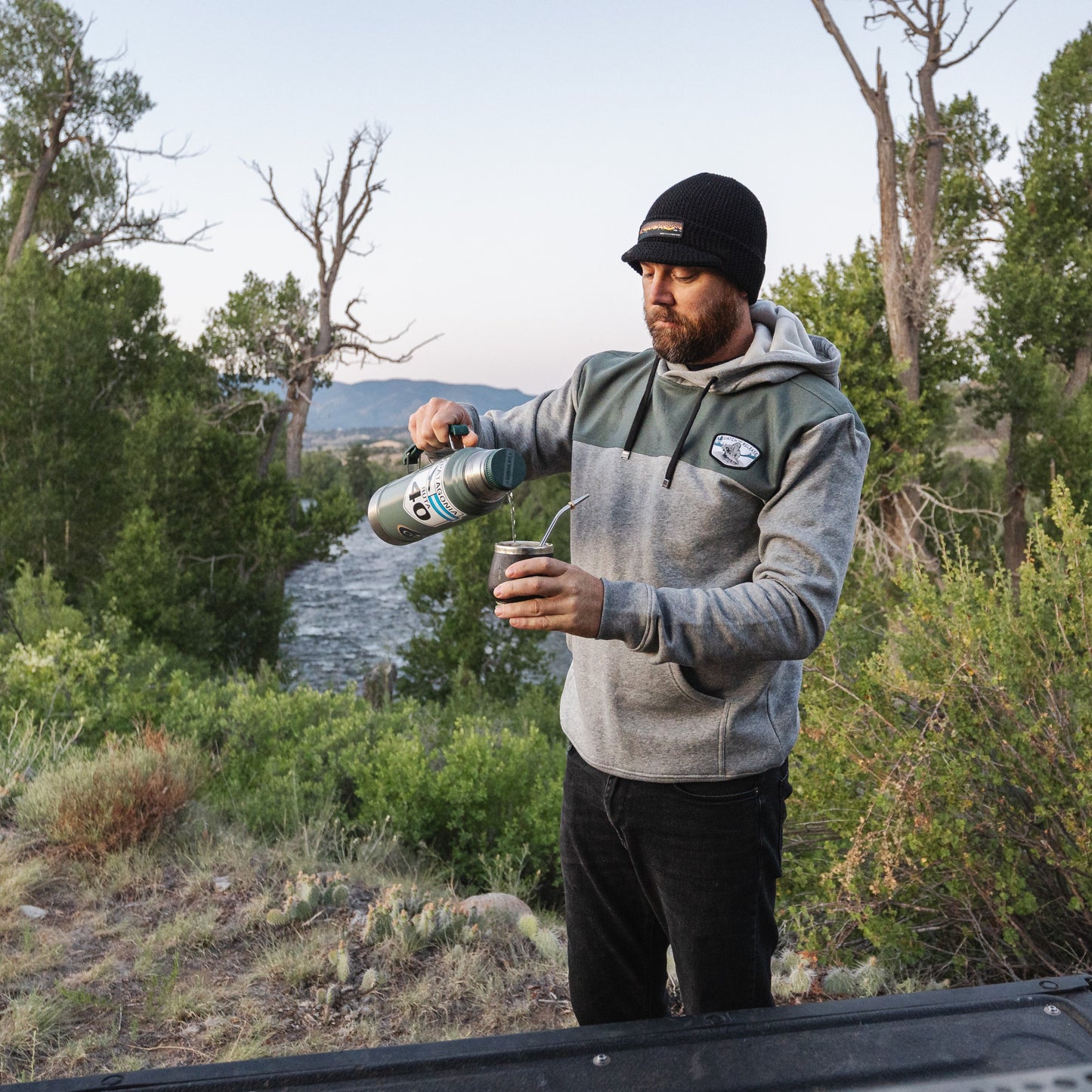 Man pouring hot coffee wearing a gray and hoodie in the mountains with a river int he background