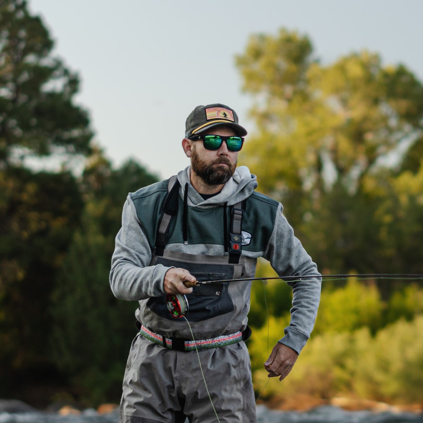 Man standing in river fishing with a green hat and gray goodie